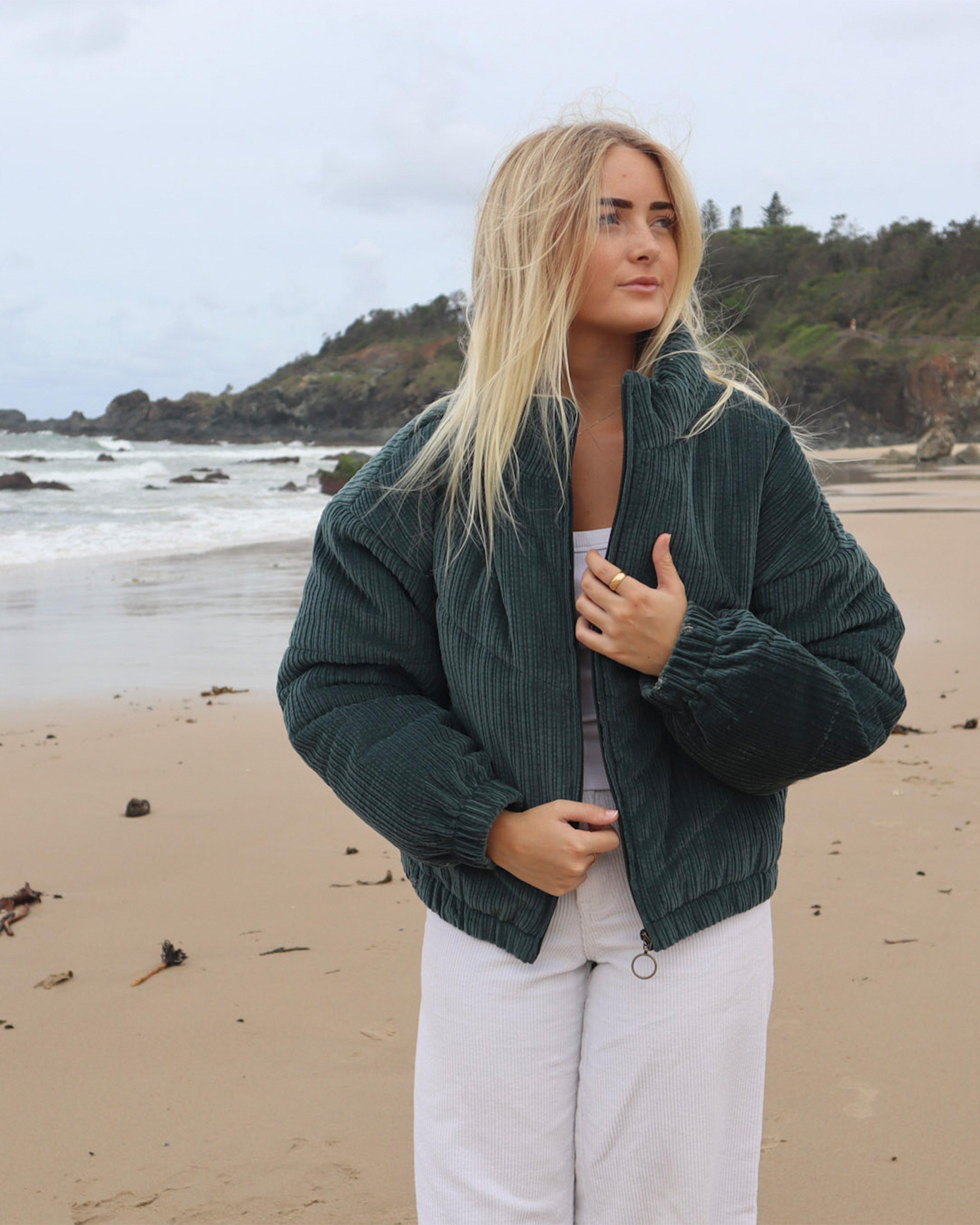 Dark green cord puffer jacket worn open over white outfit on the beach