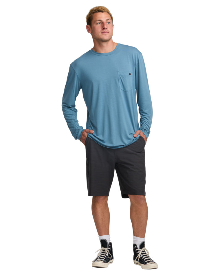 Full body view of man wearing black shorts and a light blue long sleeve tee with hands in pockets.