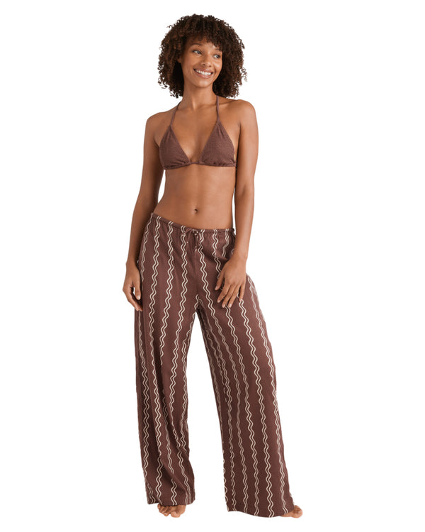 Woman wearing brown printed wide-leg pants and a matching brown triangle bikini top, smiling with one hand in pocket.