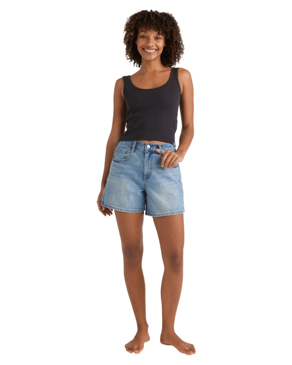Woman wearing a black tank top and mid-blue high-waisted denim shorts, standing barefoot, smiling.