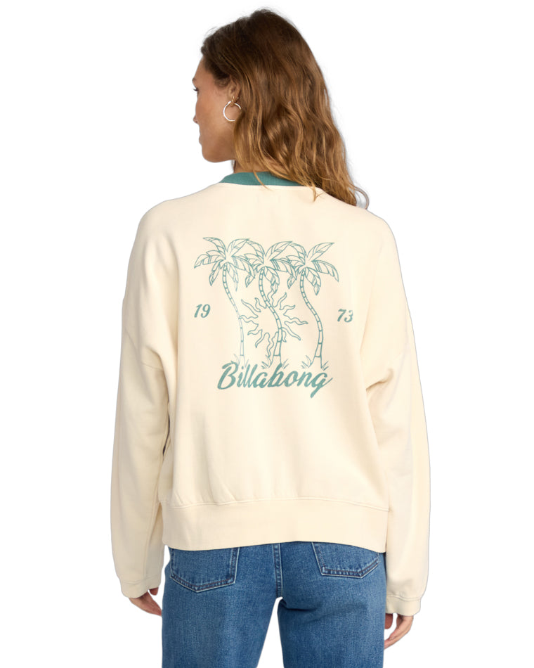 Cream crew neck sweatshirt featuring a teal Billabong graphic, shown from a side angle with one hand in the pocket.