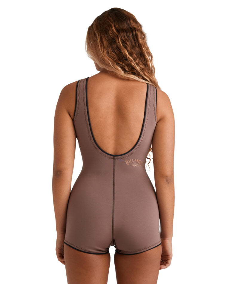Woman wearing a sleeveless brown wetsuit with black trim, standing front-on with arms behind her back.