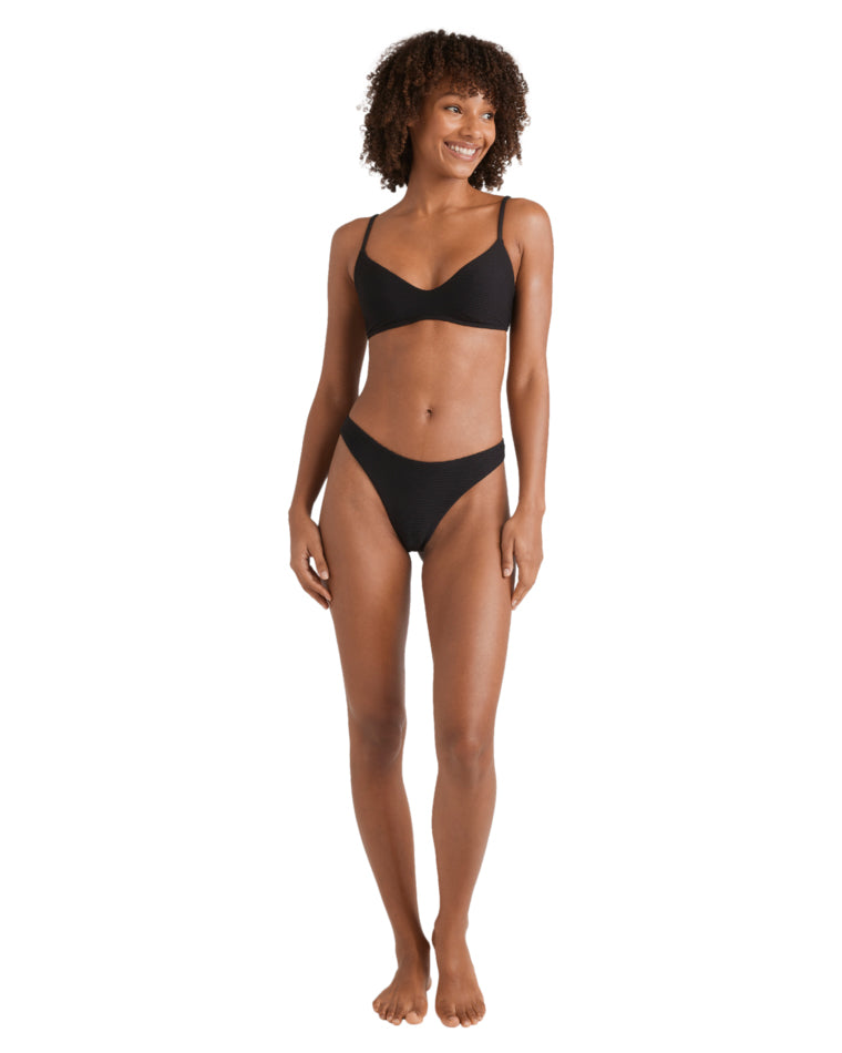 Full body view of a woman in a black bikini with ribbed texture and thin straps, standing barefoot.