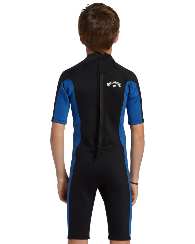 Boy wearing a black and blue Billabong short sleeve wetsuit, back view with zipper and Billabong logo.