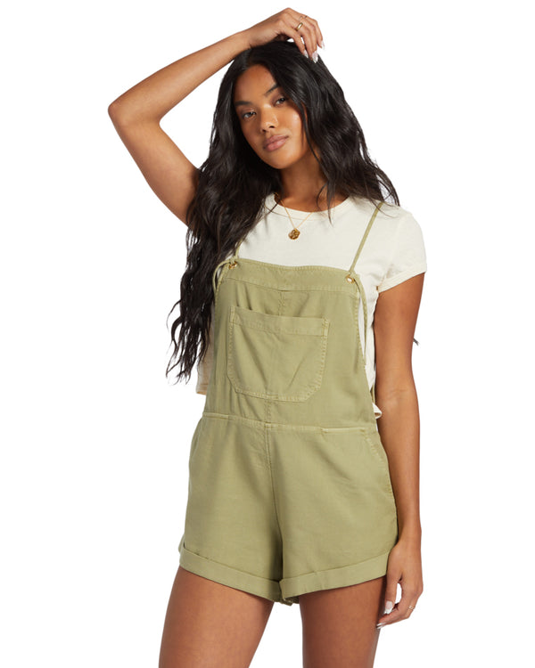 Woman wearing olive green short overalls with a front pocket and rolled hems, styled over a white tee.