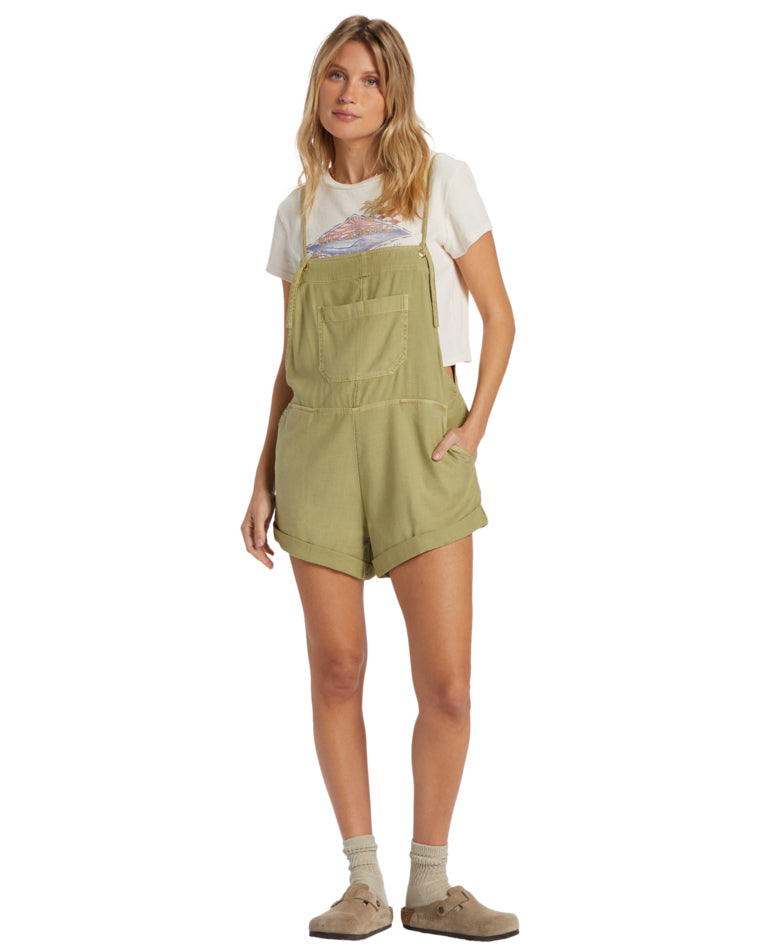 Woman in olive green short overalls layered over a cropped white tee, styled with socks and clogs.