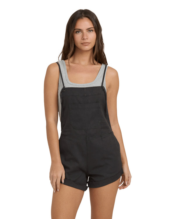 Woman front view wearing black short overalls with a grey scoop neck tank underneath, standing against a plain white background.