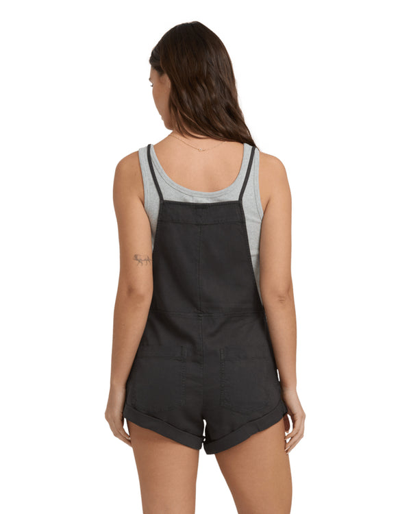 Woman back view wearing black short overalls and a grey scoop neck tank, standing against a plain white background.
