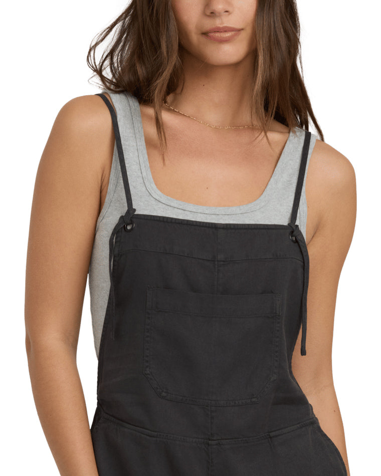 Close-up front view of a woman wearing black short overalls with a grey scoop neck tank.
