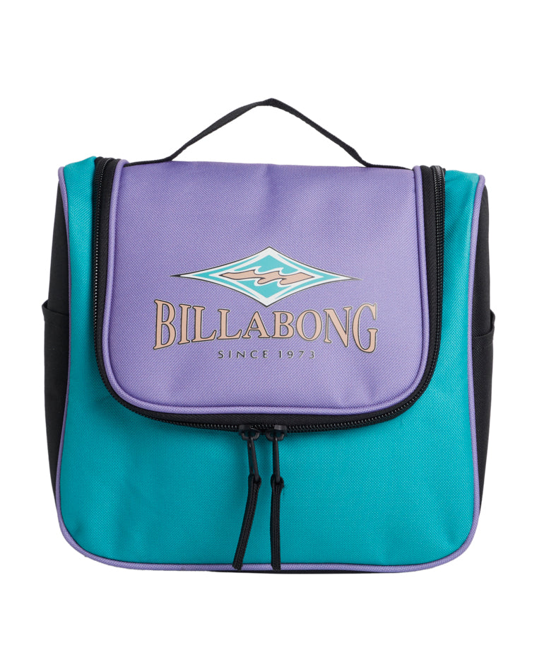 Front view of a Billabong travel bag featuring a purple flap with logo and "Since 1973" text, teal body, black sides, and dual zip closure.
