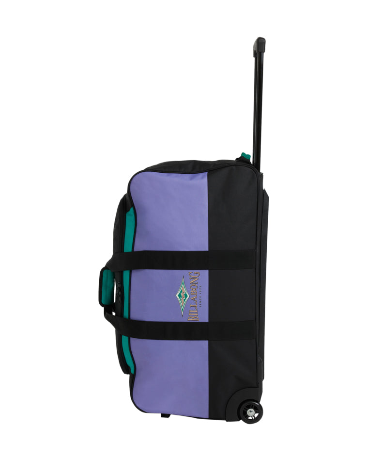 Side view of a large Billabong wheeled travel bag in a colour-blocked design featuring purple, black, and green panels, with logo printed on the front and extended pull handle upright.