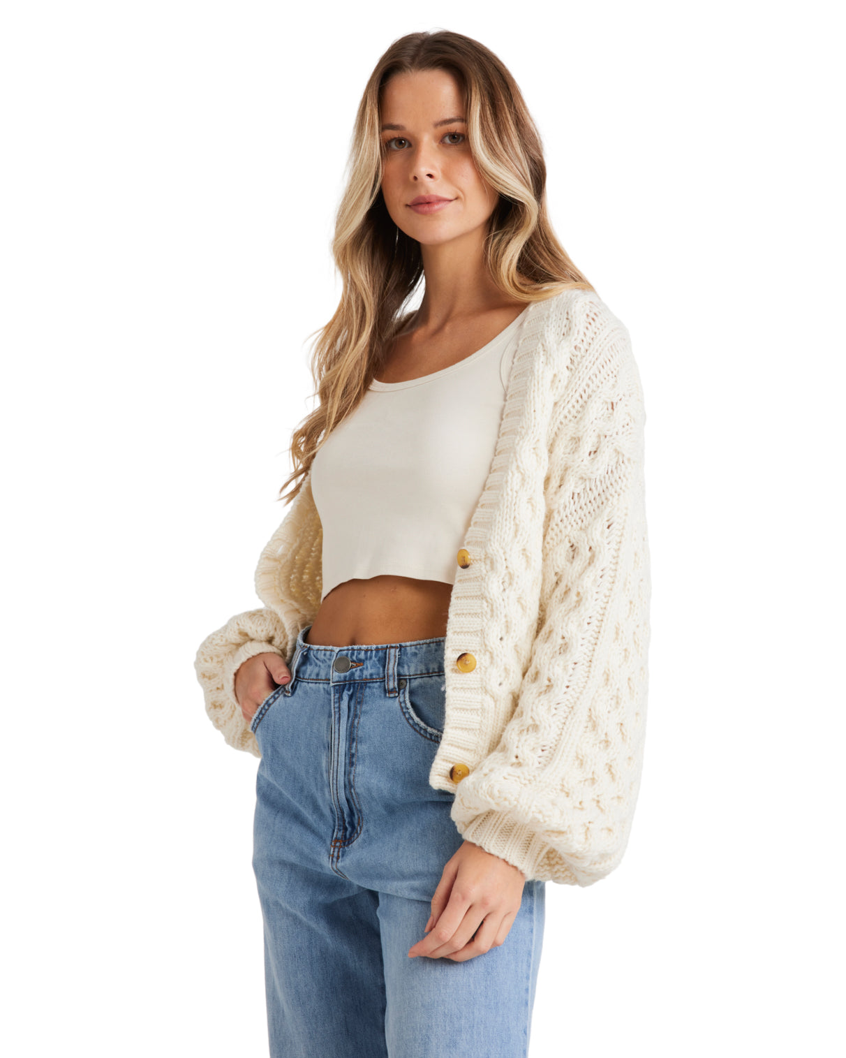 Female model wearing a cream chunky knit cardigan with wooden buttons, a white crop top, and high-waisted blue jeans.