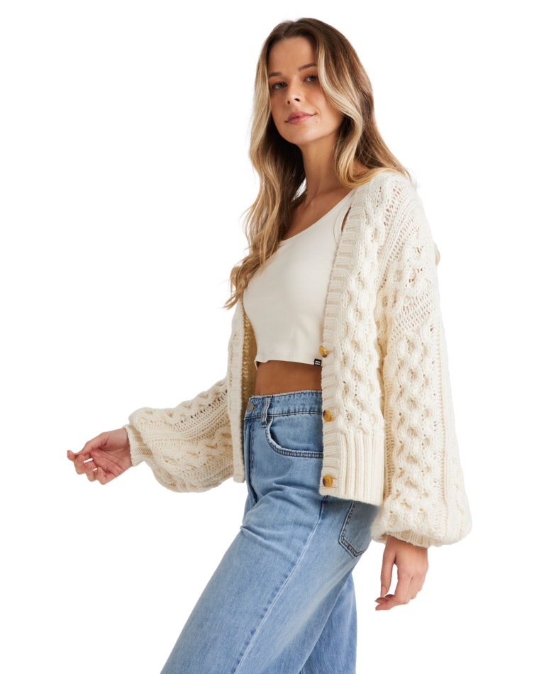 Side view of a female model wearing a cream chunky knit cardigan with cable patterns, balloon sleeves, and button details.
