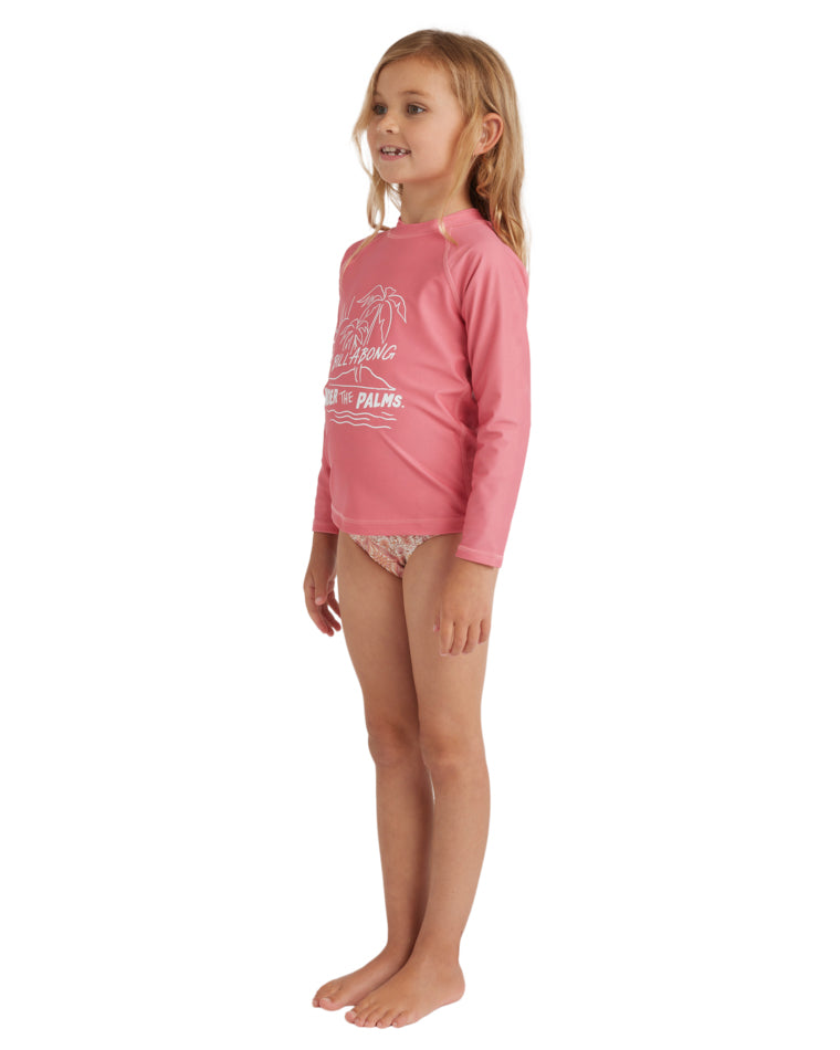 Young girl side-front view wearing a pink long sleeve Billabong rash vest with “Under the Palms” print and floral bikini bottoms.
