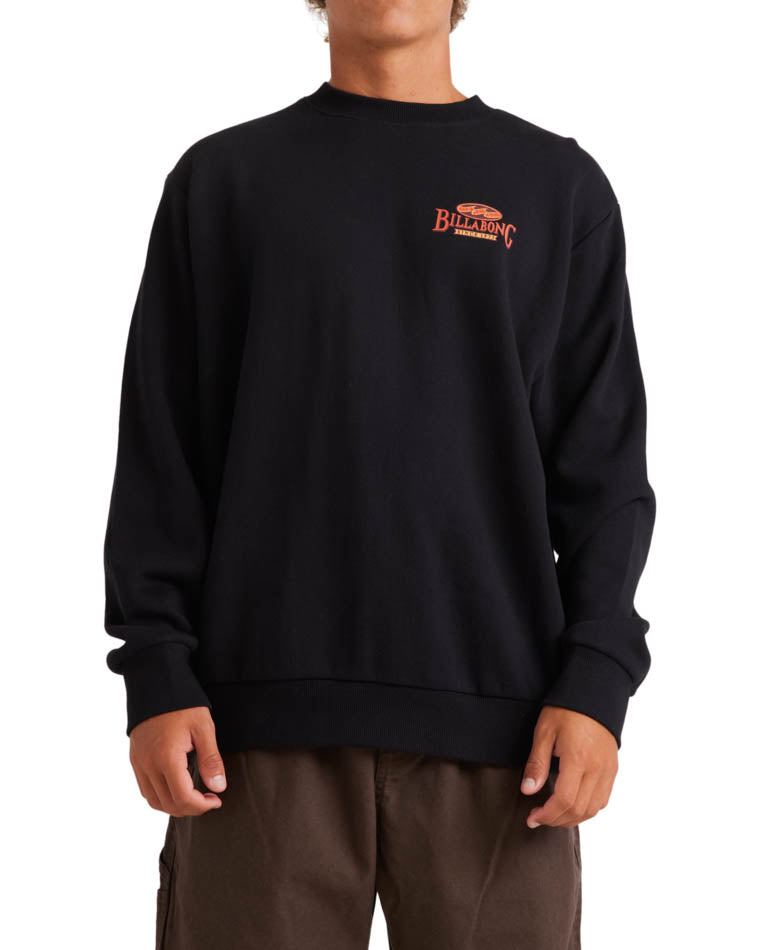 Model wearing a black Billabong crewneck sweatshirt with a small red logo on the left chest, paired with brown pants.