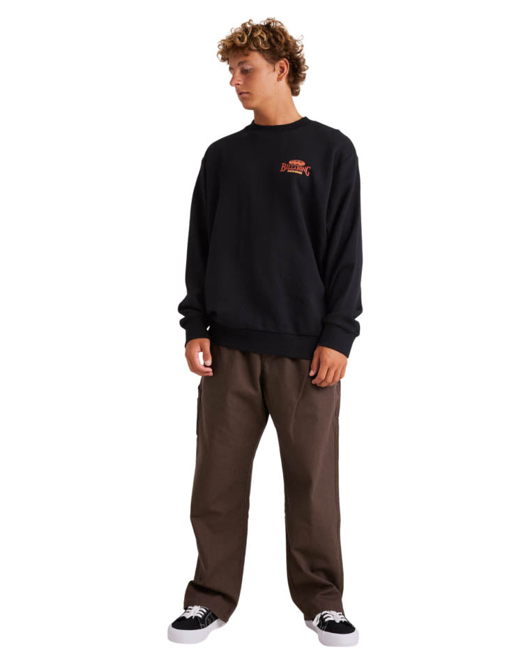 Full-body shot of a model wearing a black Billabong crewneck sweatshirt, brown pants, and black-and-white sneakers.