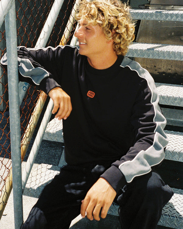 Man sitting on metal stairs, wearing a black crewneck sweater with grey contrast piping on the sleeves and a small chest logo.