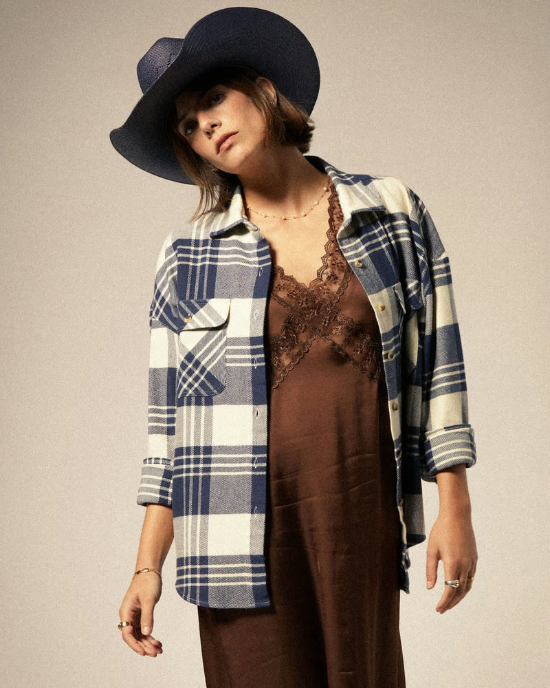 Woman wearing an oversized blue and white plaid flannel shirt layered over a brown lace-trimmed slip dress and cowboy hat.