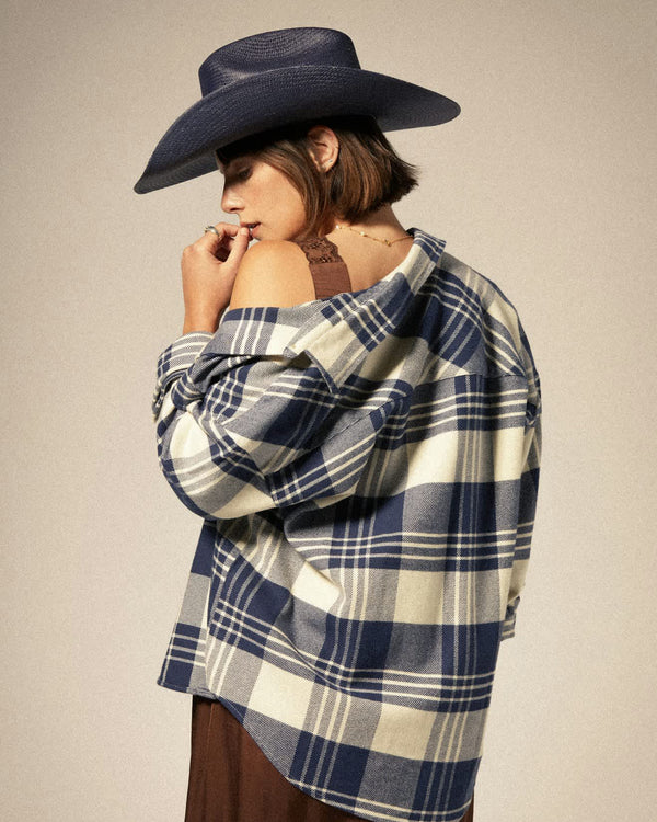 Woman wearing an oversized blue and white plaid flannel shirt draped over her shoulders, paired with a cowboy hat and dress.