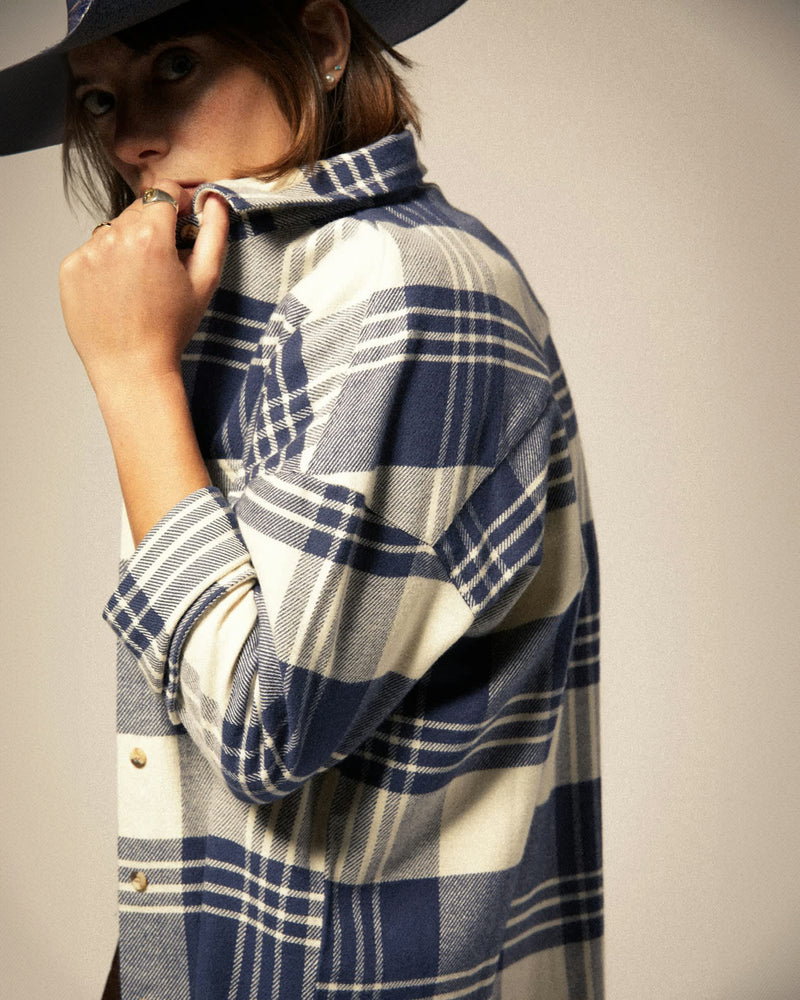 Woman wearing a blue and white plaid flannel shirt with gold buttons, pulling the collar up while accessorized with rings.