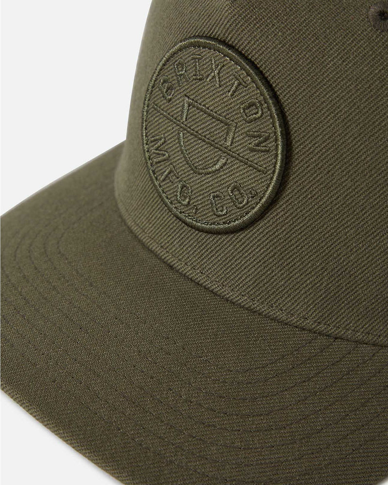 Close-up view of an olive green cap with an embroidered circular Brixton logo on the front panel and detailed stitching.