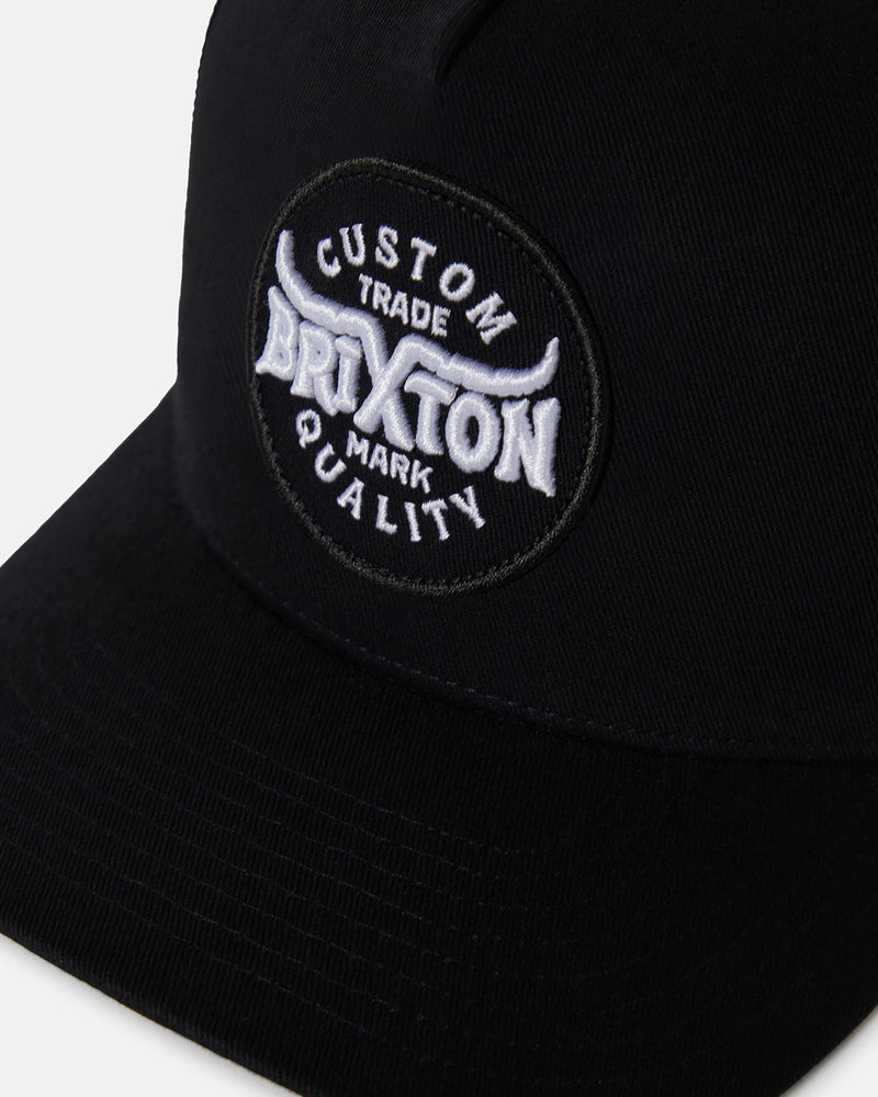 Close-up of a black cap featuring an embroidered patch with "Custom Brixton Trade Mark Quality" text in white and black.