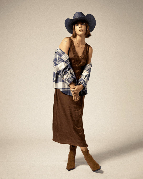 Elegant brown satin slip dress with delicate lace trim, styled with a plaid shirt, cowboy hat, and suede ankle boots.