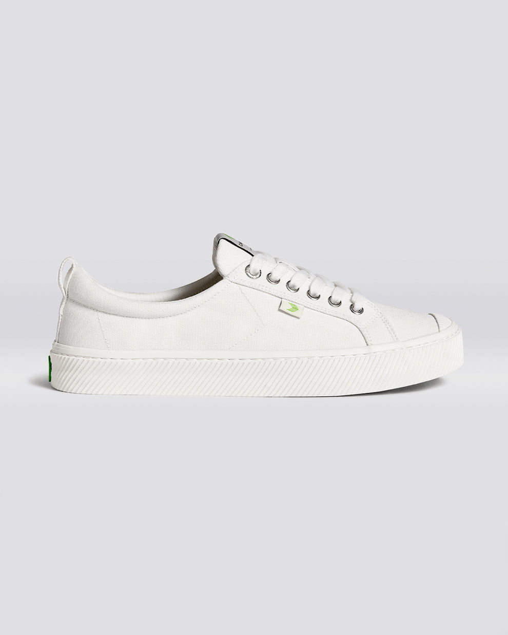 A side view of white low-top sneakers featuring a textured canvas upper, silver eyelets, white laces, and a green logo detail.