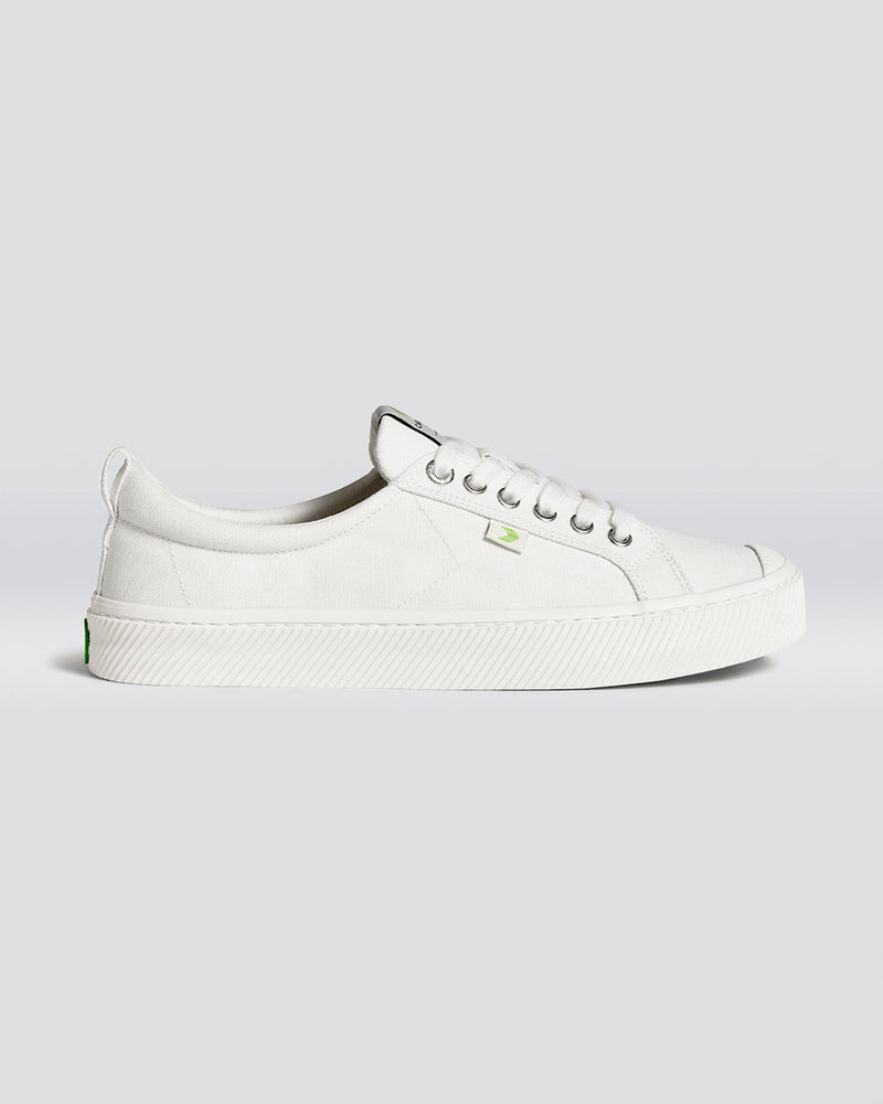 A side view of white low-top sneakers featuring a textured canvas upper, silver eyelets, white laces, and a green logo detail.