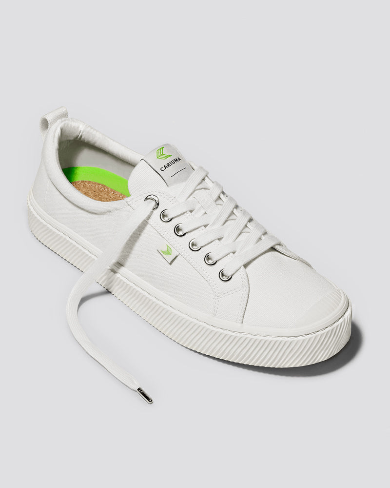 A white low-top sneaker with silver eyelets, green branding on the tongue and side, a cork insole, and textured rubber soles.