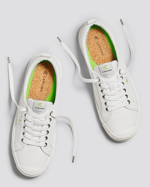 A pair of white low-top sneakers with silver eyelets, green branding, cork insoles, and textured rubber soles, shown from above.