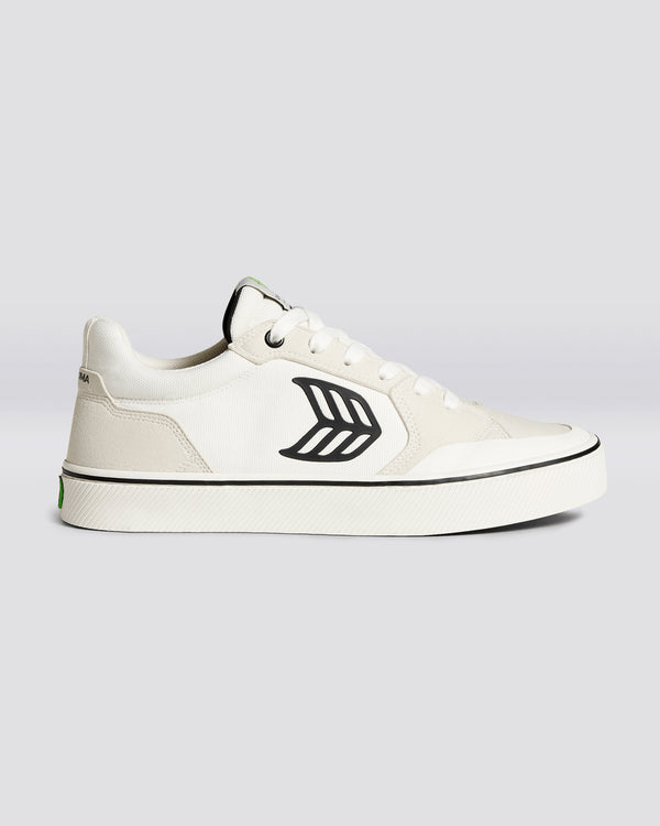 White low-top sneaker with black leaf logo, lace-up closure, textured rubber sole, and subtle green branding on heel.