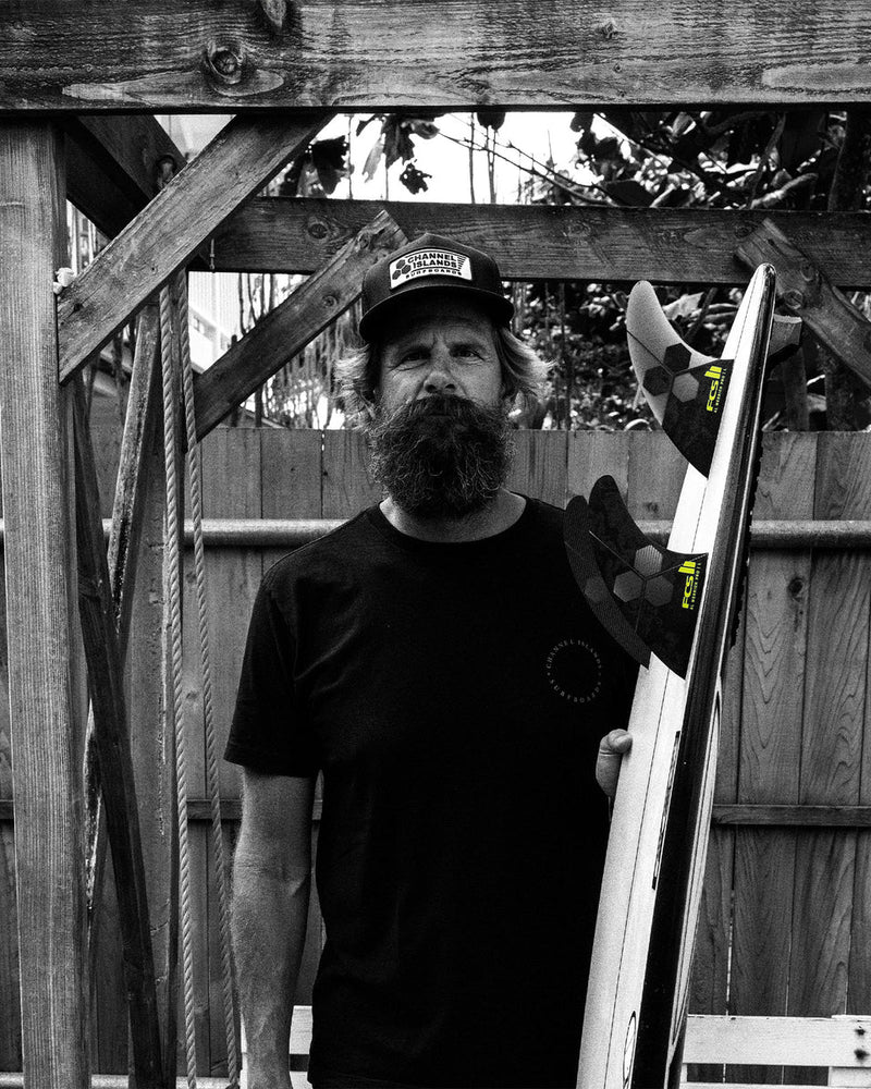 Bearded man in black tee holding a surfboard with FCS II Al Merrick Pro fins