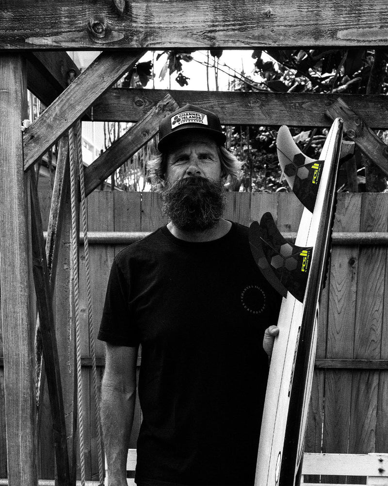 Bearded man in black tee holding surfboard with FCS II Al Merrick Pro fins in a timber outdoor setting