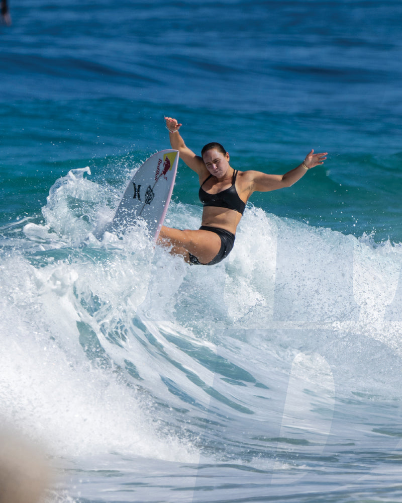 Female surfer performing a top turn on a wave wearing black swimwear with ocean spray around her board