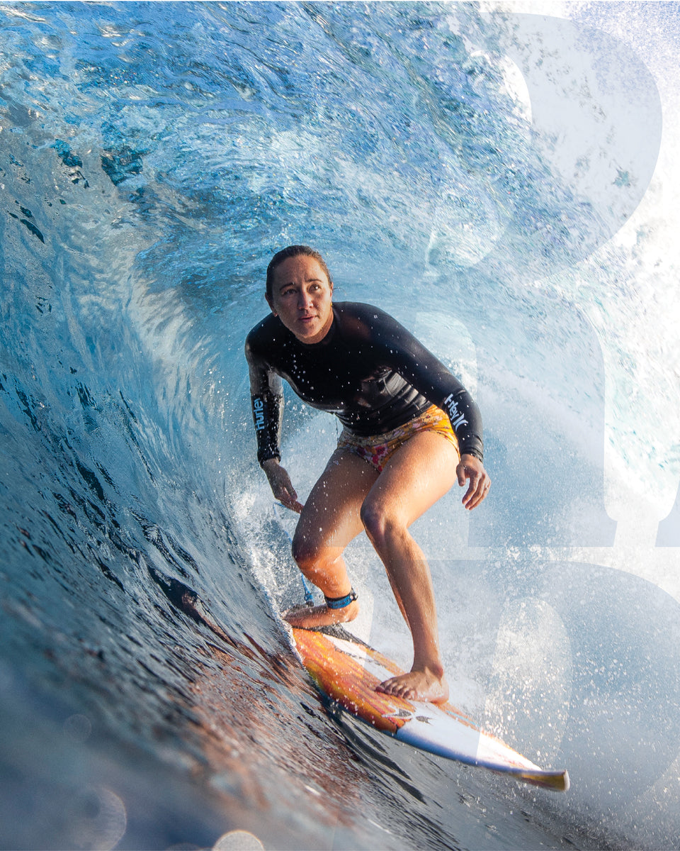Female surfer riding inside a barrel wave on an orange and white board, wearing a black rashie and floral shorts.