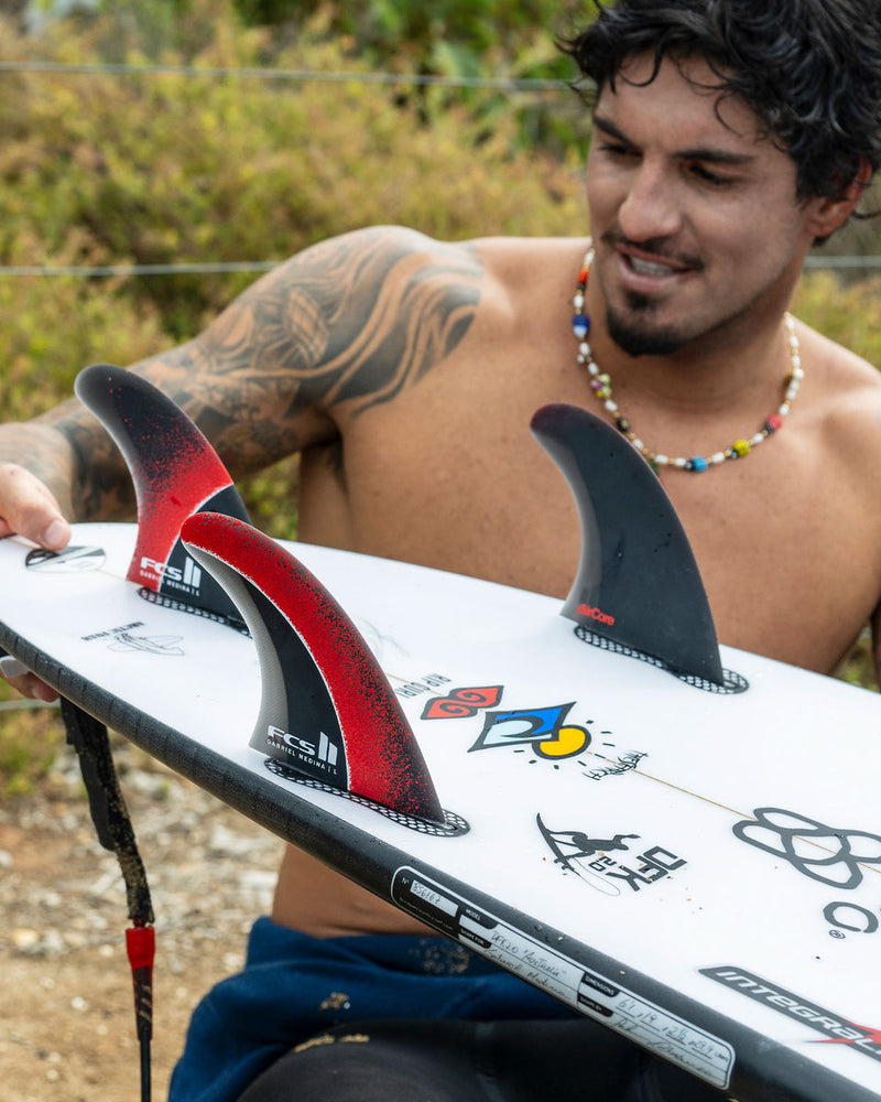 Surfer checking FCS II Gabriel Medina fins setup on a white shortboard with custom decals.
