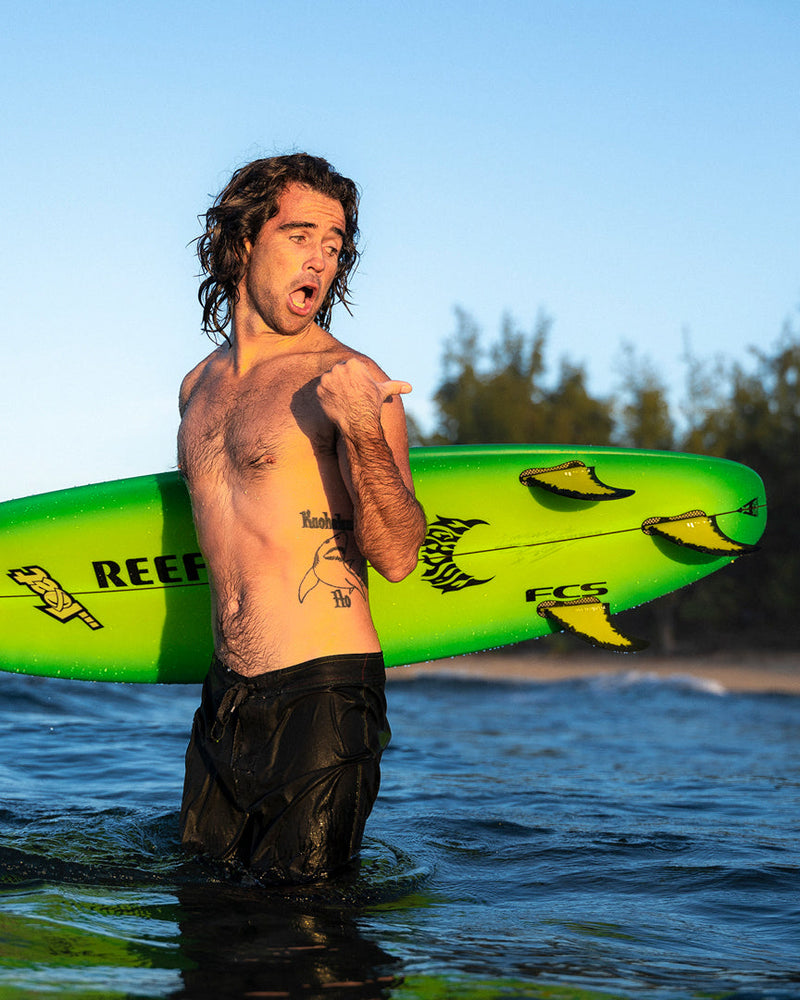Surfer holding green board fitted with Mason Ho fins, standing in the ocean at golden hour.