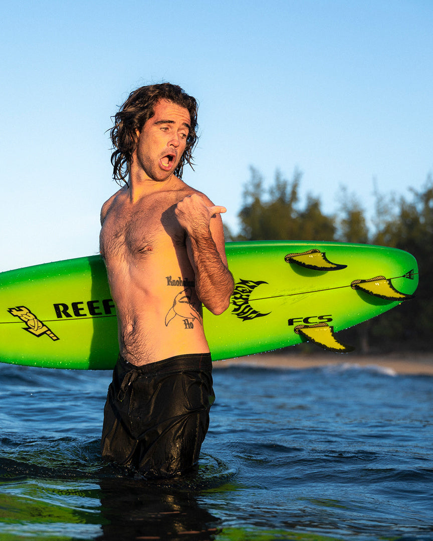 Surfer posing in the ocean with a green board fitted with yellow Mason Ho fins and bold black graphics.
