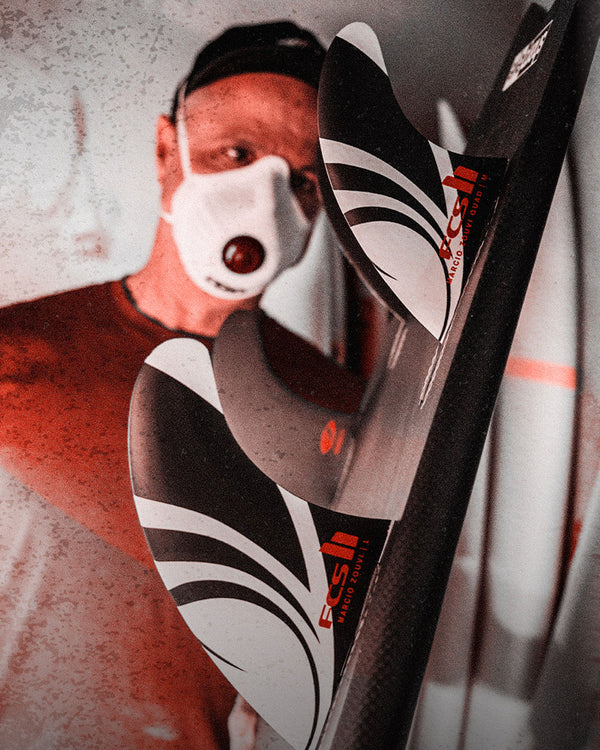 Man inspecting surfboard setup with FCS II Marcio Zouvi fins, wearing a mask in a shaping bay.