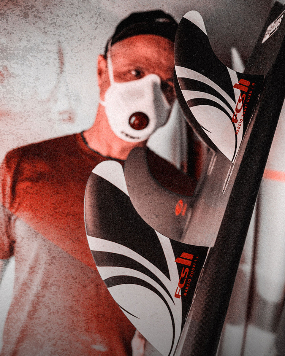 Man wearing a mask inspecting a surfboard fitted with FCS II Marcio Zouvi fins in a shaping bay.