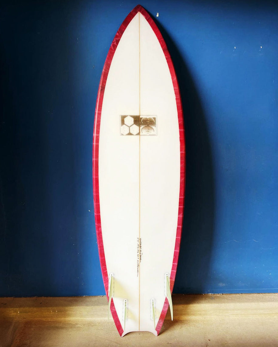 Channel Islands surfboard with red rail tint, white deck, quad fin setup and black hexagonal logo, shown upright.
