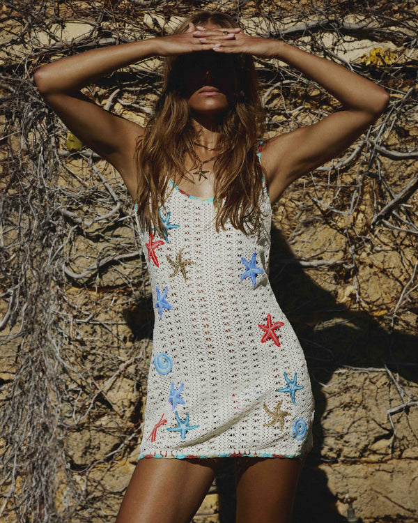 Model wearing a crochet mini dress with colorful starfish and seashell embroidery, posing against a textured rocky background.