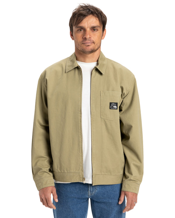 Man wearing a khaki Quiksilver zip-up jacket with a front pocket and logo patch, paired with a white t-shirt and jeans.