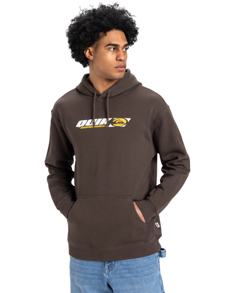 Man wearing a brown Quiksilver hoodie with a retro logo on the chest, paired with light blue jeans, standing casually.