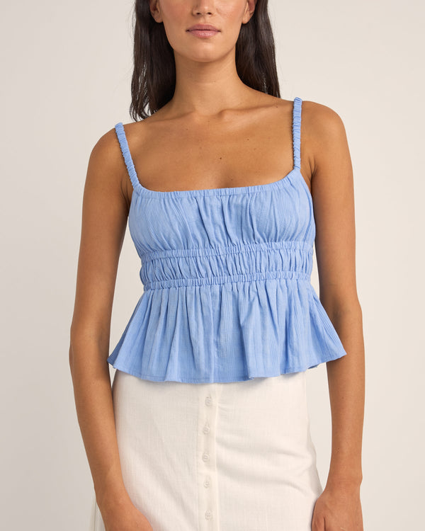 Light blue sleeveless top with ruched detailing, elasticated straps, and a flowy peplum hem, paired with a white skirt.