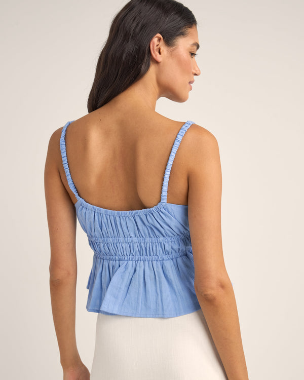 Back view of a light blue sleeveless top with ruched detailing, elasticated straps, and a flowy peplum hem, paired with a skirt.