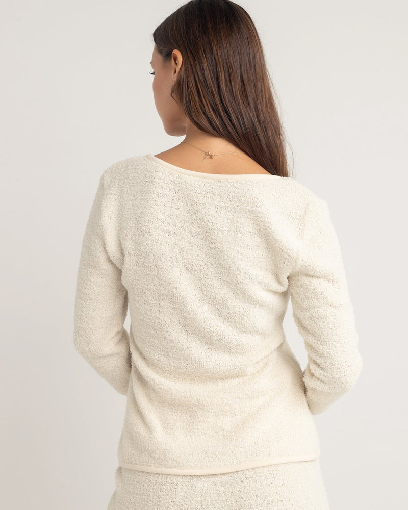 Back view of a cream-colored textured long-sleeve top with a wide neckline and fitted silhouette, paired with a skirt.