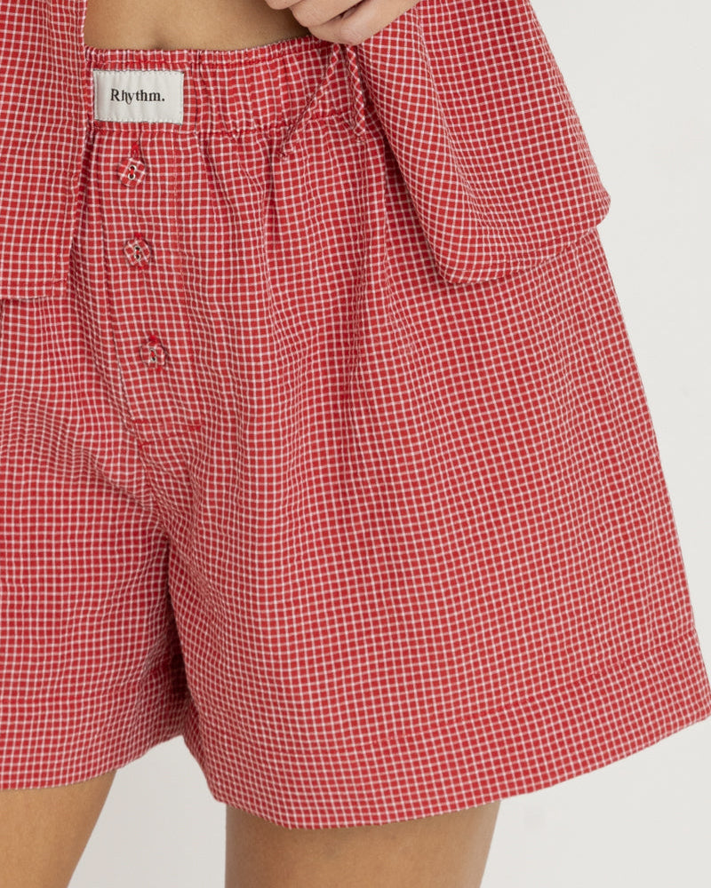 Close-up of red gingham shorts featuring an elastic waistband, button details, and a Rhythm logo tag on the front.