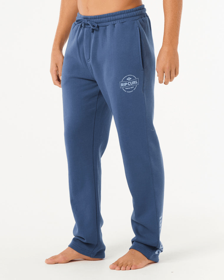 Side view of man wearing blue Rip Curl sweatpants with elastic waistband, drawstring, and logo print on upper leg.